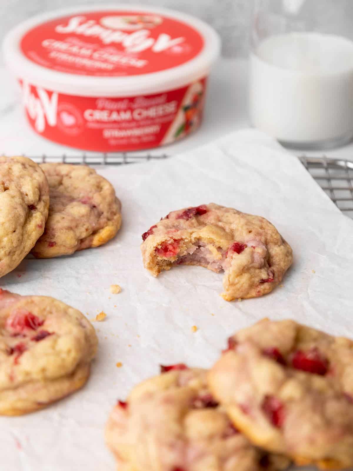 A strawberry cheesecake cookie has been bit into, showing the texture and cream cheese filling inside. It is surrounded by more cookies, a milk jar, and SimplyV's strawberry cream cheese.