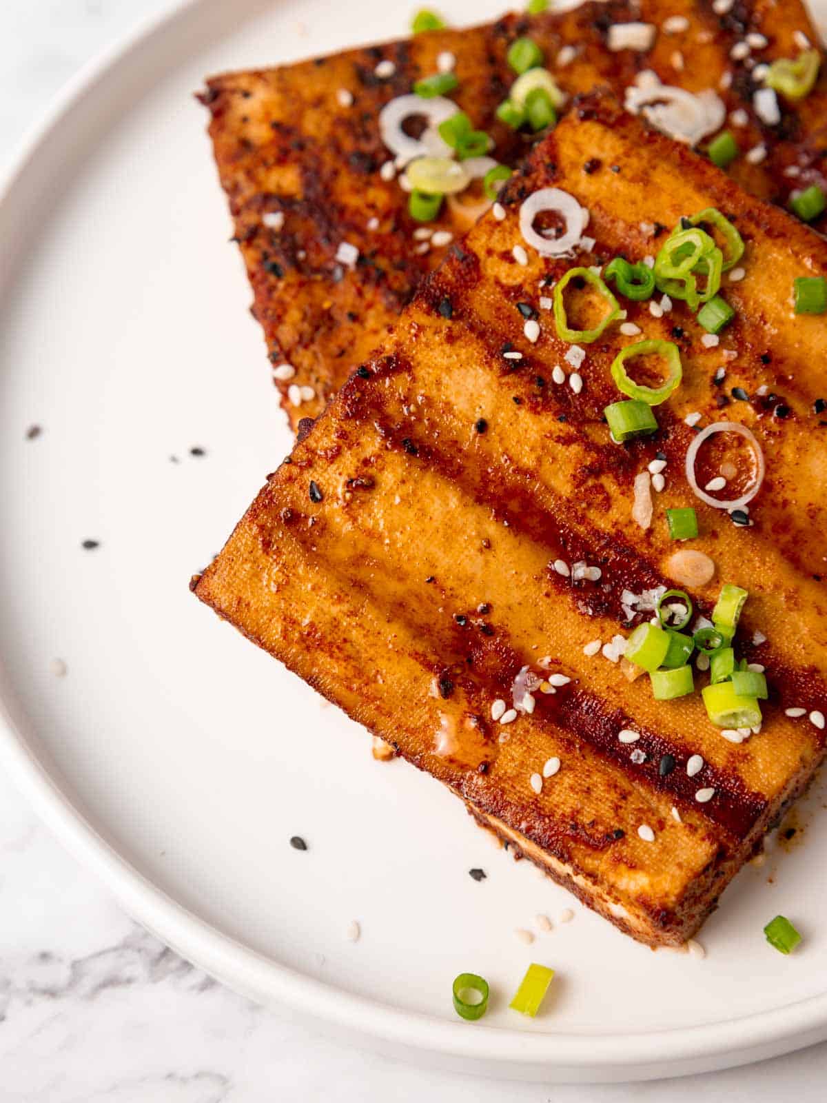 Juicy grilled tofu steaks garnished with scallions and sesame seeds served on a plate.