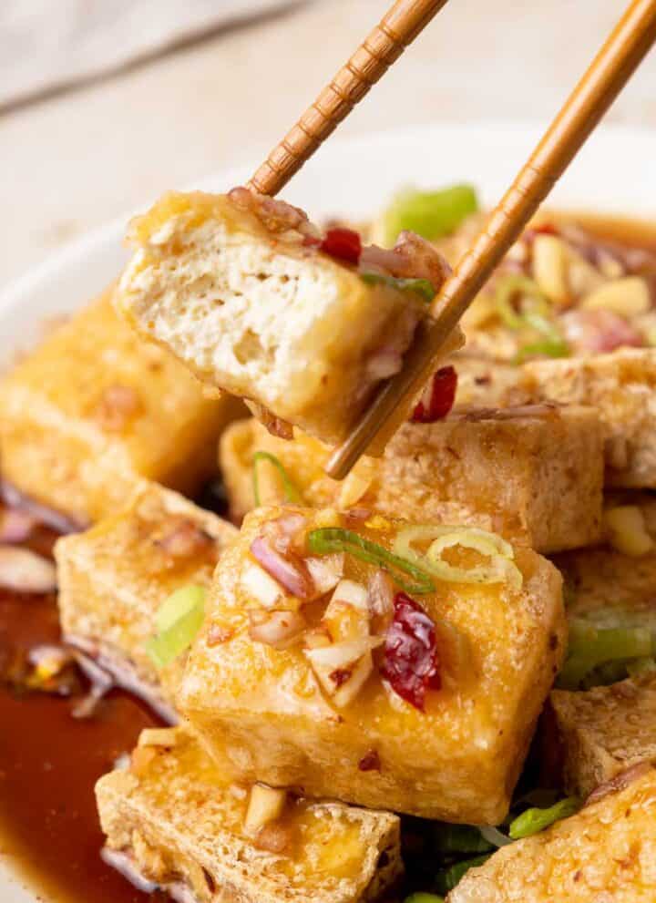 Tahu Gejrot served on a plate. A Tofu piece is being grabbed by chopsticks. It is bitten into showing the fluffy texture inside.