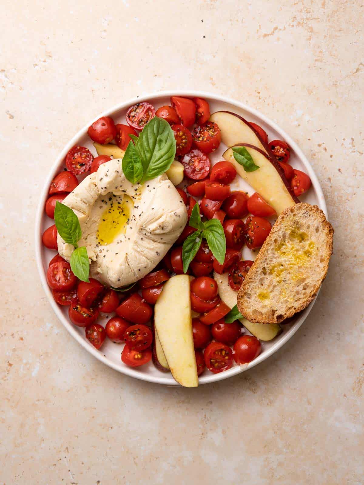 Vegan Burrata served on a round plate as a Caprese salad with cherry tomato, drizzles of olive oil, fresh basil, artisan bread, salt, pepper, and apple slices.