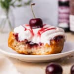 Cherry donut that has been bit into is topped with whipped cream and cherry jam served on a plate.